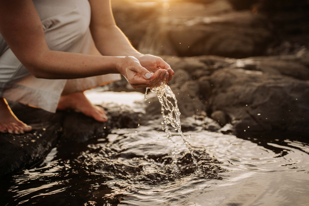 A person in a white sleeveless top kneels by a rocky stream at sunset, cupping water that splashes into the pool. The background is a blurred natural outdoor setting with warm golden light.